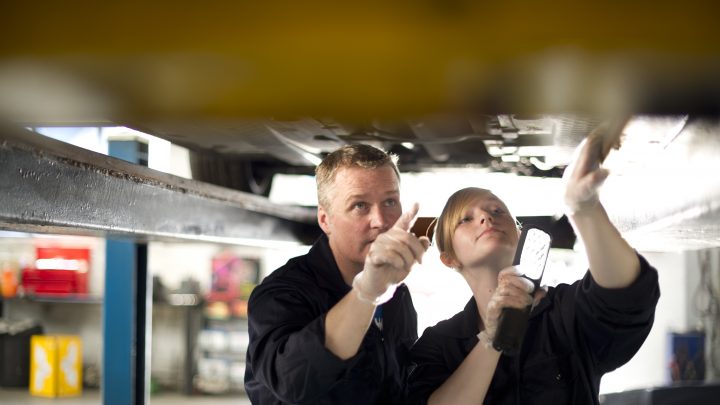 Eine Auszubildende zur Mechanikerin steht unter einem Auto zusammen mit ihrem Ausbilder, der ihr etwas erklärt.