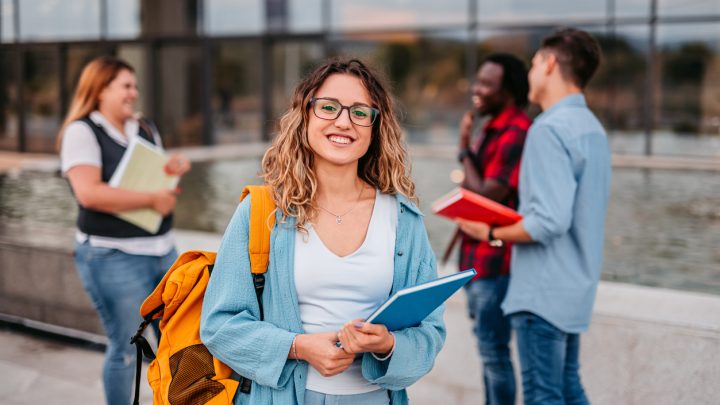 Eine lächelnde Studentin steht mit ihrem Rucksack und einem Buch auf dem Campus. Im Hintergrund unterhalten sich ihre Freunde miteinander.